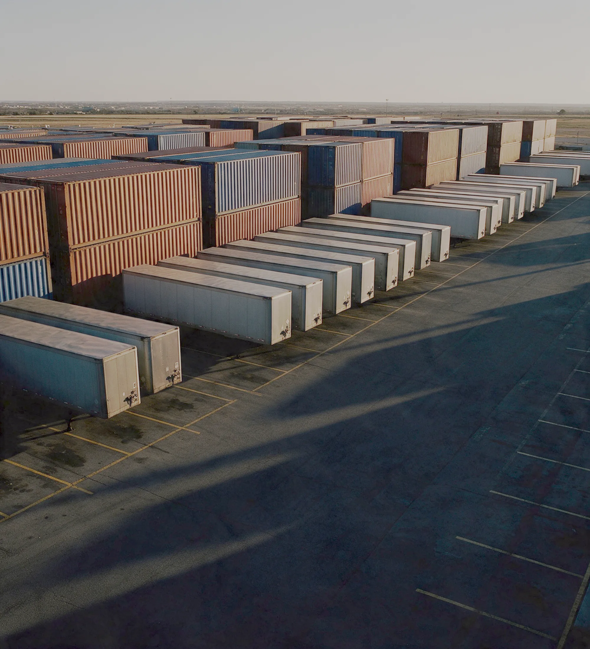 Rows of shipping containers and trailers on the asphalt of a freight yard in late afternoon light.