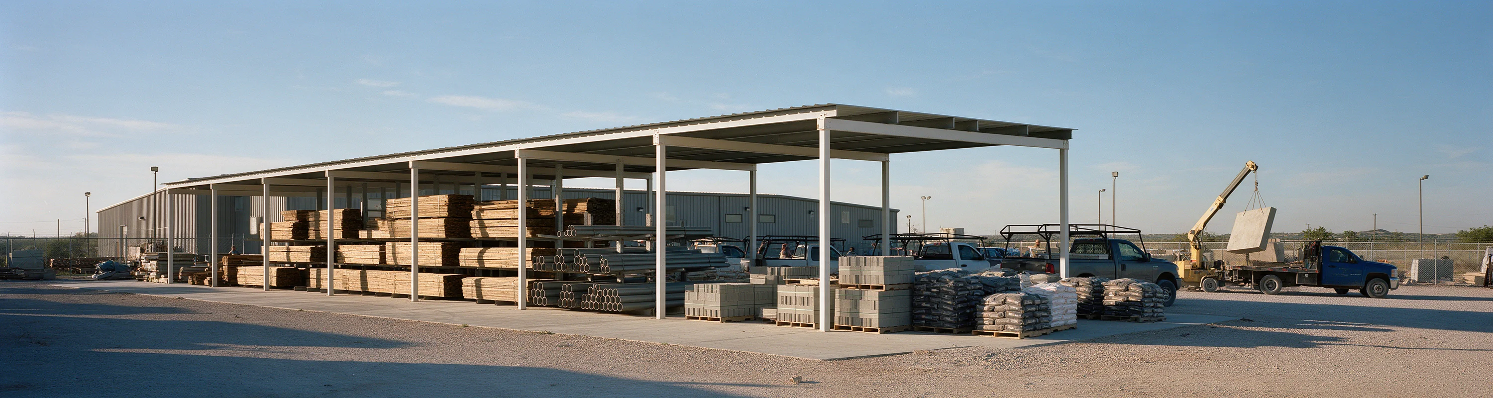 A contractor supply yard in the morning, lumber and pipe racked under a long steel canopy.