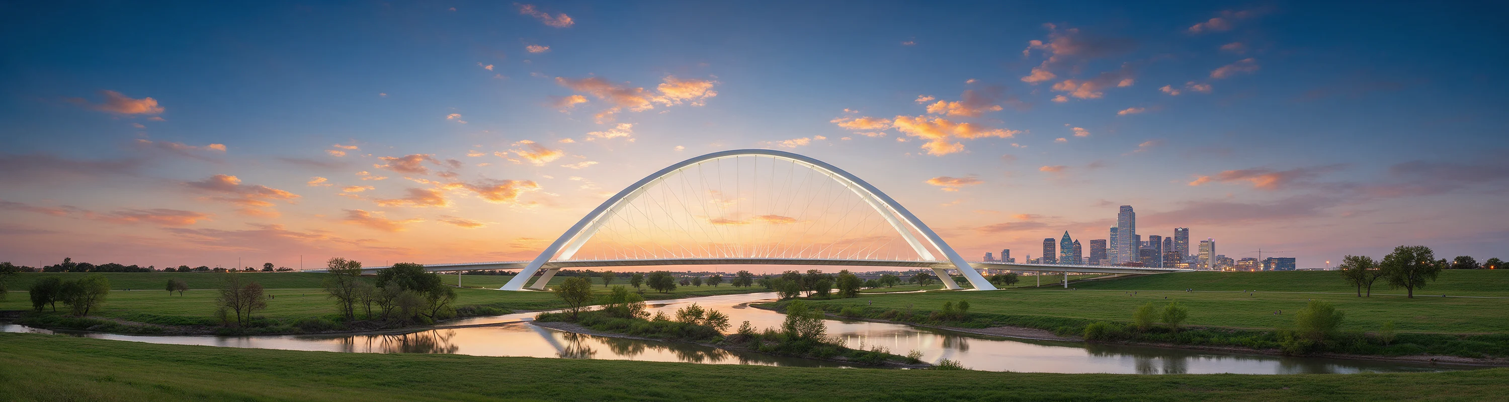 A white arch bridge over river flats at sunset, the Dallas skyline beyond it.